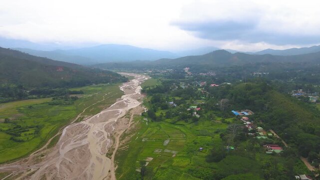 A vast aerial landscape view of a river valley in rural Aileu, Timor-Leste. The winding riverbed cuts through green plains and farmland, surrounded by hills and mountains.