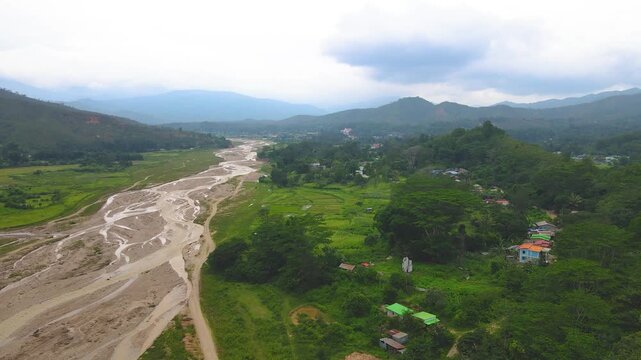 A vast aerial landscape view of a river valley in rural Aileu, Timor-Leste. The winding riverbed cuts through green plains and farmland, surrounded by hills and mountains.