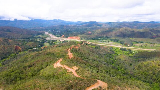 A vast aerial landscape view of rural Aileu, Timor-Leste. A red dirt road cuts through the green and brown hills, with a river winding through the valley below distant mountains.
