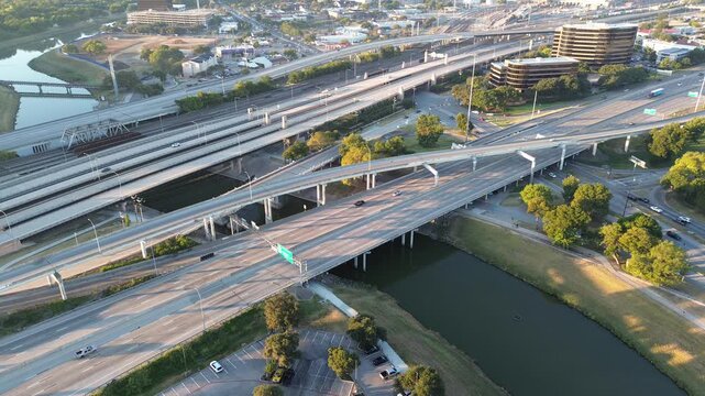 Busy highway traffic and early morning runners, walkers trace Trinity Trails beside river, elevated channel toward Fort Worth hospital district. Green buffers soften the city infrastructure edge