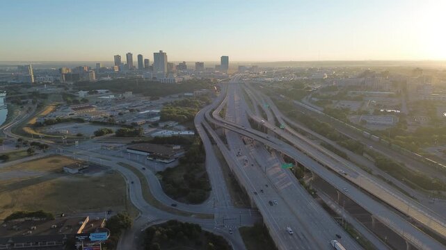 Fort Worth four-level stack interchange near downtown, with I-30 and I-35W converging beside the Trinity River. Sunrise light reveals layered ramps, skyline silhouettes, busy commuter flow, Texas