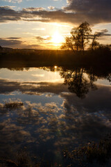 Vibrant sunset sky reflecting in peaceful river, silhouetted trees along the bank with golden light. Picturesque nature scene