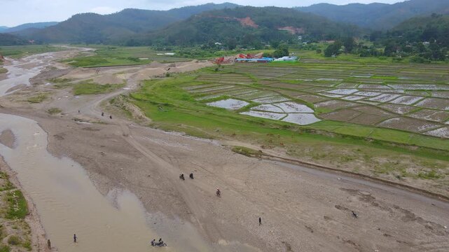 Motorcyclists cross a wide, dry riverbed used as a makeshift road in rural Aileu, Timor-Leste. A scene highlighting poor infrastructure and accessibility challenges.