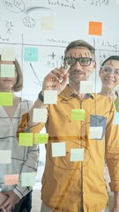 Professional team brainstorming, sharing strategy on glass whiteboard with colorful sticky notes, working collaboratively in bright office space