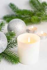 Festive holiday ambiance with lit glowing candles in glass jar, green fir tree branches and silver shiny christmas ornament baubles on white wooden table prepared for cozy winter celebration party