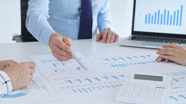 Business professional pointing at printed charts with a pen during a meeting, laptop showing bar graphs and calculator on table while team collaborates on financial analysis and planning