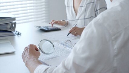 Two professional accountants working together, using a magnifying glass and a calculators, to audit financial reports in their brightly lit office. Audit and taxes in business