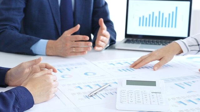 Business people reviewing financial charts and reports in office, using a laptop and calculator while engaging in discussions about strategy and performance