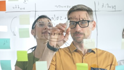 Professional team brainstorming project strategy, placing colorful sticky notes on glass board, analyzing data, sharing insights, developing collaborative solutions