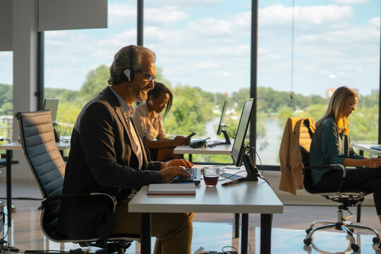 Diverse business professionals wearing headphones, typing on computers, and using mobile phones while providing customer service in a bright, contemporary open-plan office setting - Powered by Adobe