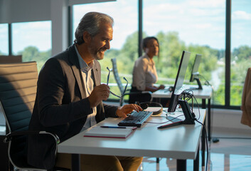 Smiling mature businessman holding glasses while typing on a keyboard at a modern office desk with a female colleague blurred in the background, focused on work and collaboration