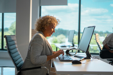 Senior businesswoman with curly hair sitting at her desk, typing on a computer keyboard and wearing...