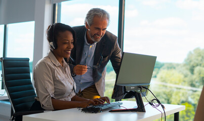 Smiling african american woman wearing a headset assisting an older male mentor at a computer screen, collaborating on customer service in a modern office