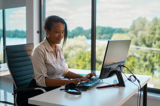Professional african american woman sitting at a desk, typing on a computer keyboard in a bright, modern office space with a large window overlooking a scenic outdoor landscape