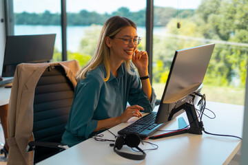 Young professional woman smiling while working at her desk on a computer, wearing a casual shirt and glasses, in a bright office with green landscape visible outside