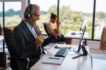 Mature call center agent wearing a headset and glasses, excitedly raising fists in a gesture of success while watching a computer screen at a spacious office desk