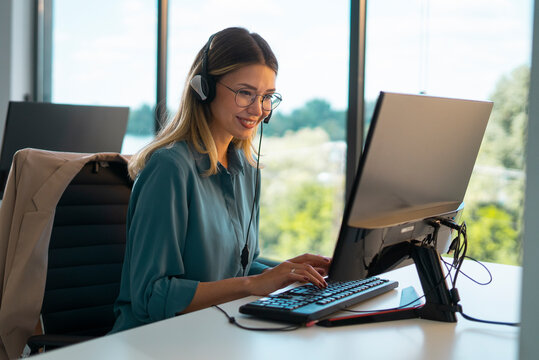 Young woman in glasses wearing a headset, smiling while typing at a computer in a modern call center, providing friendly customer support and remote assistance from her desk - Powered by Adobe