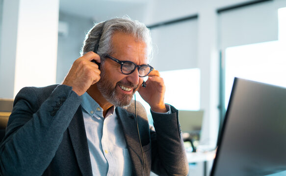 Mature man with a salt and pepper beard and glasses smiling confidently, wearing a headset with a microphone, and engaging in a customer service call or virtual meeting at an office desk