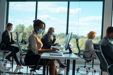 Multiracial business people wearing protective face masks and maintaining social distance while working on computers in a modern office during the coronavirus pandemic
