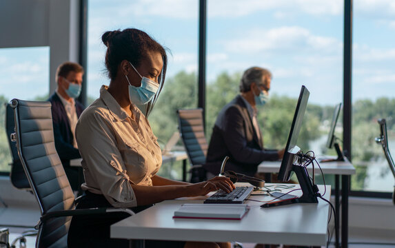 Multiracial business professionals are socially distancing and wearing protective face masks while working on computers in a modern corporate office during the covid-19 pandemic