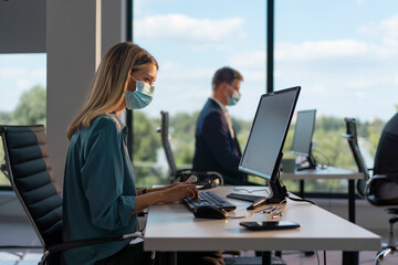 Businesswoman wearing a protective face mask while typing on a computer keyboard at her desk, with a male colleague working in the background, focusing on safety at the workplace