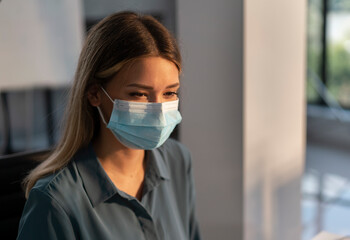 Woman wearing a surgical face mask for protection against viruses and pollution while working in a modern office, adapting to new health and safety protocols during the pandemic