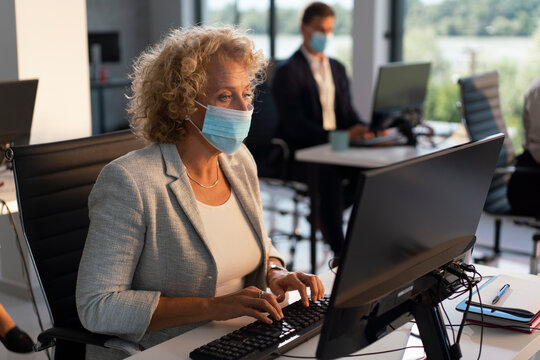 Businesswoman wearing protective face mask working at computer in modern office, typing at desk and adapting to workplace safety measures during the covid-19 pandemic - Powered by Adobe