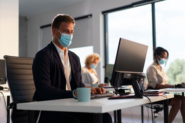 Businesspeople collaborating in a modern office, typing on computers, and wearing protective face masks, demonstrating health and safety protocols for the pandemic