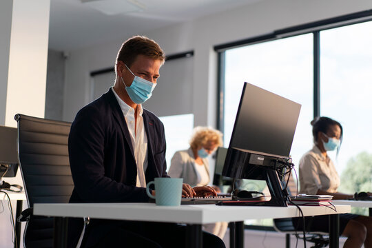 Business people working in a modern office, wearing protective face masks to ensure safety and health during a pandemic, focusing on their computers at individual desks