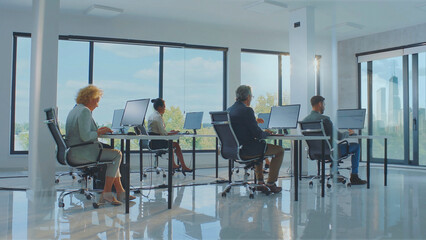 Diverse group of professionals concentrating on their desktop computers at individual workstations in a contemporary open-plan office, surrounded by large windows overlooking a city skyline