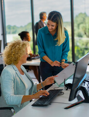 Young businesswoman smiles while presenting a clipboard to a seated senior colleague who types and...