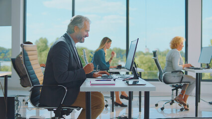 Diverse business people working on desktop computers at their desks in a contemporary open-plan office building with large windows overlooking green surroundings