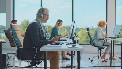 Diverse customer service representatives wearing headsets and typing on computers, providing technical support or sales assistance in a modern, brightly lit open-plan call center office environment
