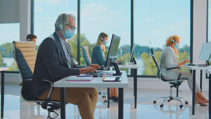 Diverse team of customer support workers wearing protective face masks and headsets, social distancing while working at computers in a modern call center office