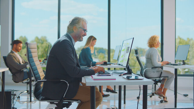 Diverse business professionals actively working on computers at individual desks in a bright, contemporary open-plan office, collaborating and focusing on their tasks during a busy workday