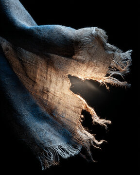 Worn fabric with frayed edges illuminated by warm light creating dramatic low key lighting effect that highlights texture and delicate threads in dark background