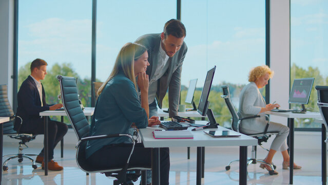 Diverse team collaborating in a bright open-plan office as a manager leans over a colleague's monitor to provide mentoring and guidance while others focus on tasks