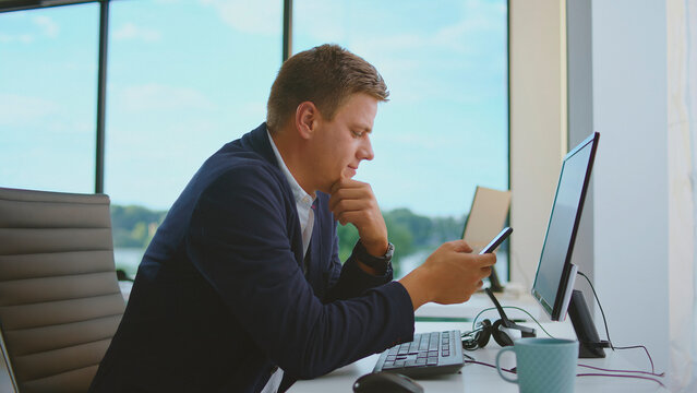 Young businessman multitasking, looking at his smartphone while sitting at his office desk, contemplating a problem or checking information with a computer nearby