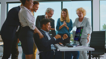 Diverse business team cheering and celebrating a successful investment and stock market victory, watching the positive results on a computer monitor in a modern office