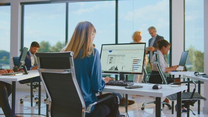 Young businesswoman working on a computer, analyzing charts and spreadsheets, while other diverse colleagues are working and casually discussing in a bright, contemporary open-plan office