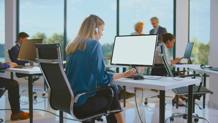 Young woman working on computer, wearing headset and typing, providing support while colleagues also collaborate in a modern, sunlit office environment
