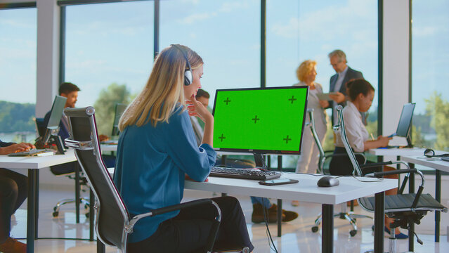 Customer service representative wearing a headset and looking at a green screen computer monitor in a modern office with diverse colleagues working in the background