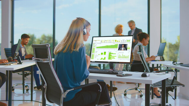 Business woman working at her desk, wearing a headset and participating in a video conference with a colleague while analyzing data on her computer in a modern office - Powered by Adobe