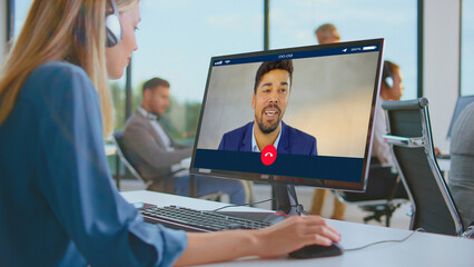 Woman wearing headphones at open-plan office desk joins video conference with male colleague on desktop screen, illustrating remote work, modern business communication and collaboration