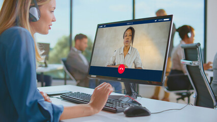 Young woman employee in headphones having a video conference call with a colleague on her computer screen, working in a modern open space office environment