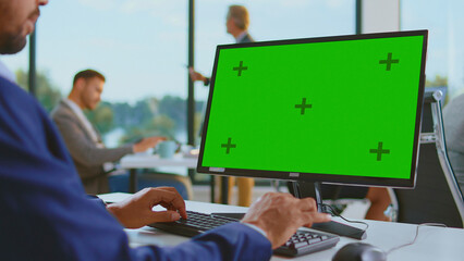 Businessman using a computer with a green screen monitor and tracking markers, typing on a keyboard while working in a modern open space office environment