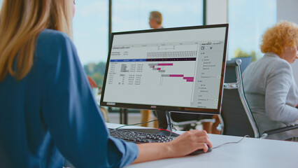 Businesswoman working on a computer screen displaying a project management gantt chart, planning tasks and tracking progress in a modern office environment with blurred colleagues in the background