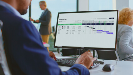 Project manager analyzing a gantt chart on a desktop monitor, planning tasks, timelines and resources in a modern office to track progress, deadlines and team workflow