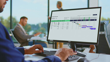 Business professional using a desktop computer to manage a project schedule and planning tasks with an organized gantt chart application in a modern office environment