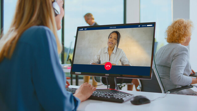 Woman in headphones sitting at her office desk and networking with a business colleague or client during an online video conference, working remotely and communicating digitally